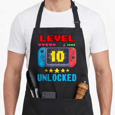 10th level birthday apron - Pixel handheld game console with LEVEL 10 UNLOCKED text, five hearts, battery icon — shown on a black cotton canvas apron against a white background