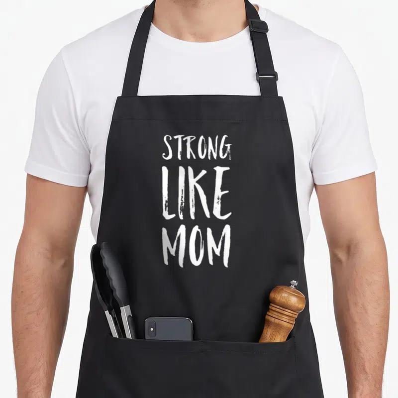 Empowered mother's day aprons - White stacked text reading STRONG LIKE MOM on dark grey background — shown on a black cotton canvas apron against a white background