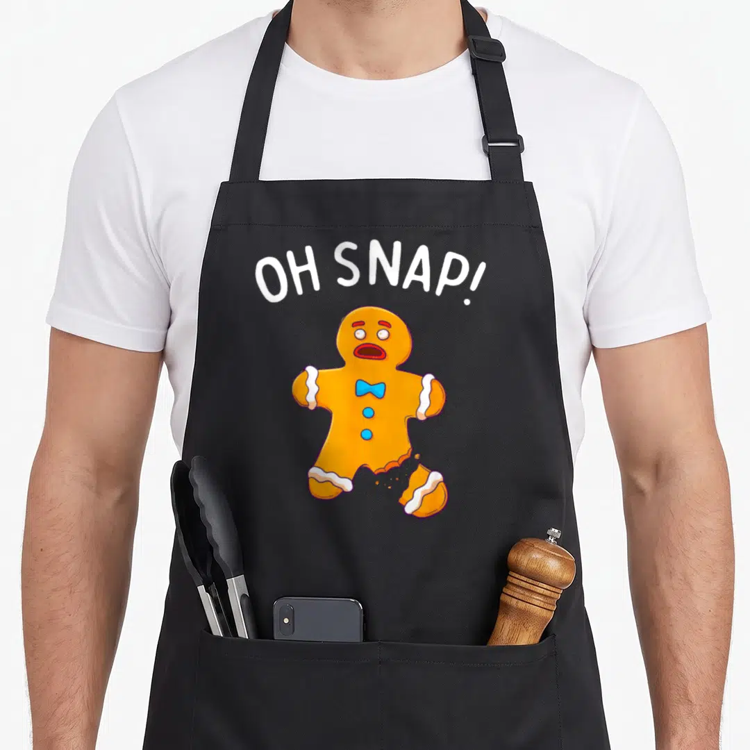 Snap gingerbread apron - Orange gingerbread man with broken leg and OH SNAP text in cartoon style — shown on a black cotton canvas apron against a white background