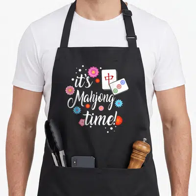 Funny chinese mahjong apron - it's Mahjong time! text with two tiles and pink blue red flowers — shown on a black cotton canvas apron against a white background