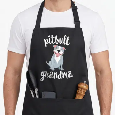 Grandparent dog apron - Cartoon grey and white pitbull sitting between white pitbull and grandma text — shown on a black cotton canvas apron against a white background