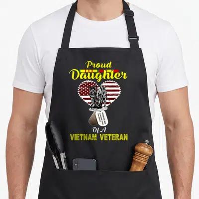 Proud wars new apron - Proud Daughter Vietnam Veteran text with soldiers in flag heart and dog tags in distressed — shown on a black cotton canvas apron against a white background