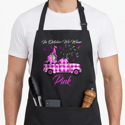Gnome breast cancer awareness aprons - Pink plaid truck gnome holding ribbon with butterflies and text In October We — shown on a black cotton canvas apron against a white background