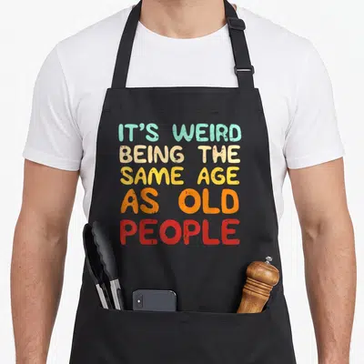 Retro general weird aprons - Stacked text IT'S WEIRD BEING THE SAME AGE AS OLD PEOPLE in teal beige yellow orange — shown on a black cotton canvas apron against a white background