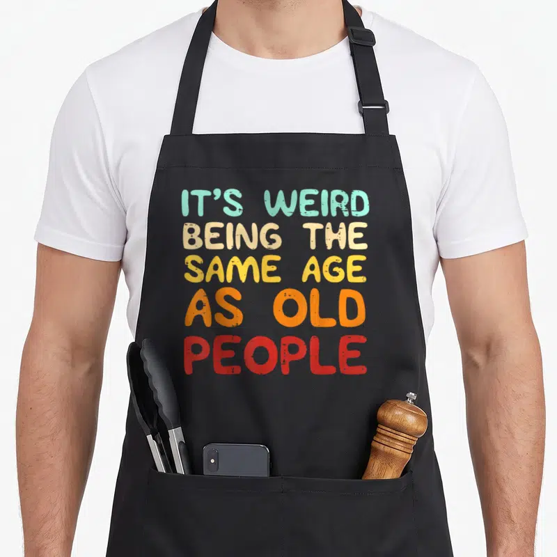 Retro general weird aprons - Stacked text IT'S WEIRD BEING THE SAME AGE AS OLD PEOPLE in teal beige yellow orange — shown on a black cotton canvas apron against a white background