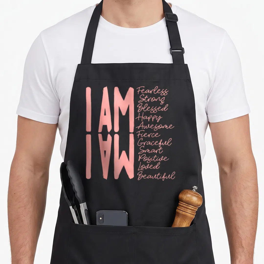 Strong fifty and fearless apron - Pink I AM typography with list of twelve positive adjectives on black fabric — shown on a black cotton canvas apron against a white background