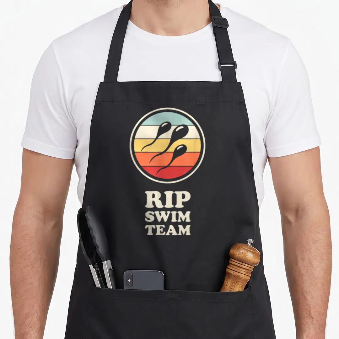 Vasectomy recovery day apron - Three grey sperm swimming across multi-colored stripes above stacked RIP SWIM TEAM text — shown on a black cotton canvas apron against a white background