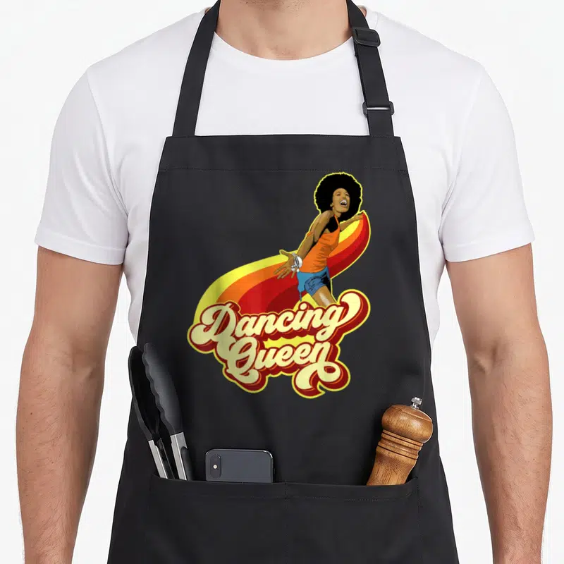 Dancing afro 70s apron - Cartoon woman with afro dancing above Dancing Queen text in yellow, red, and orange swooshes — shown on a black cotton canvas apron against a white background