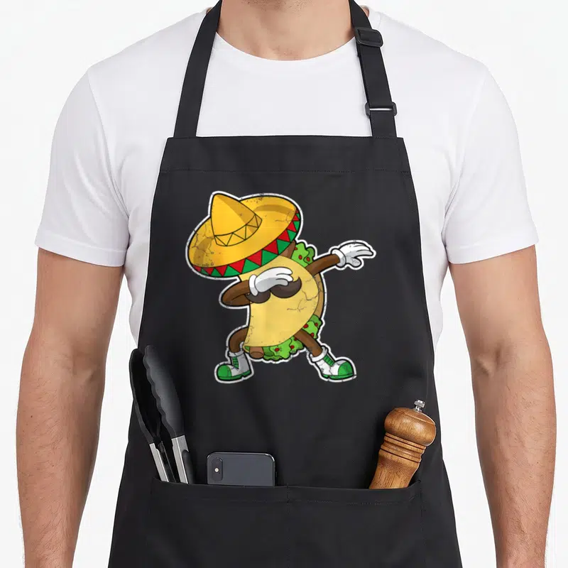 Dabbing mexico apron - Cartoon taco character in sombrero and shoes dabbing in yellow, green, red, and brown — shown on a black cotton canvas apron against a white background