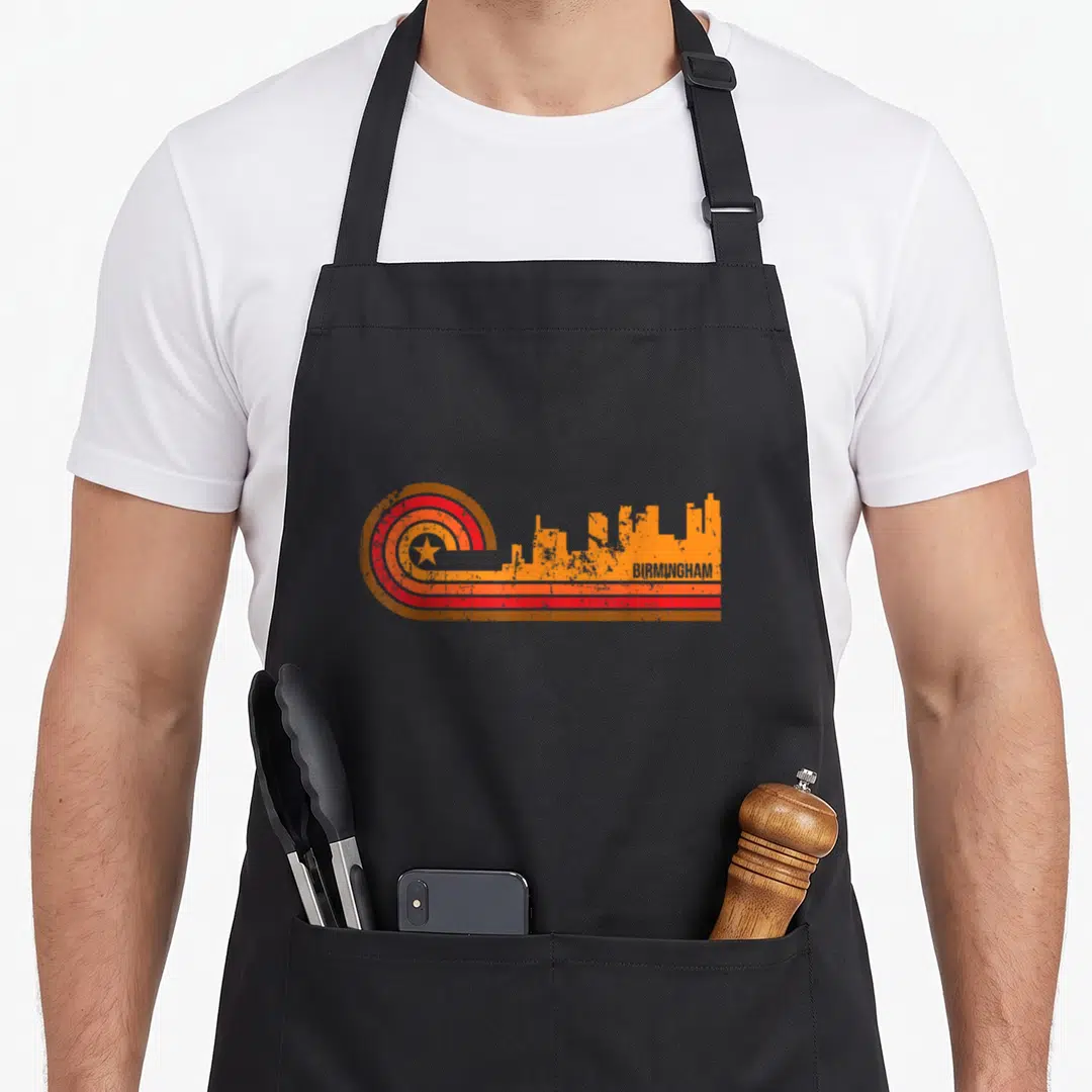 Skyline birmingham apron - Retro orange skyline with red star circles and BIRMINGHAM text — shown on a black cotton canvas apron against a white background