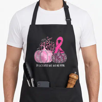 Pumpkin ribbon apron - Three patterned pumpkins and pink ribbon with text IN OCTOBER WE WEAR PINK — shown on a black cotton canvas apron against a white background