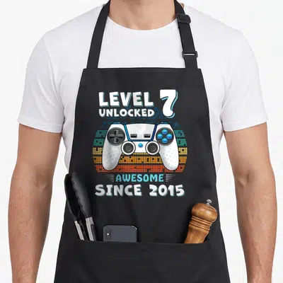 7yr dad and son aprons - Game controller graphic with LEVEL 7 UNLOCKED AWESOME SINCE 2015 text in cartoon style — shown on a black cotton canvas apron against a white background