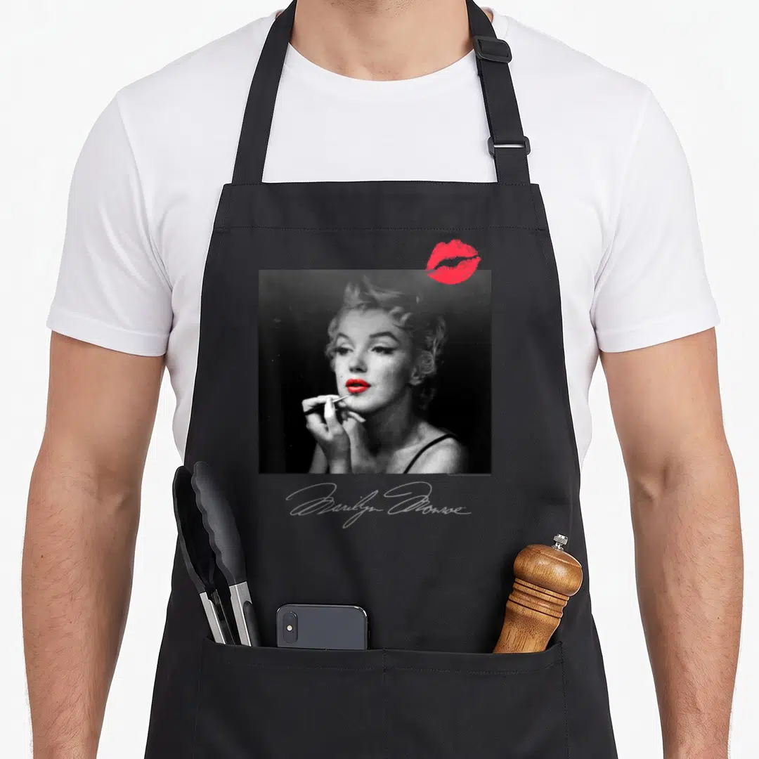 Marilyn makeup apron - Grayscale Marilyn Monroe photo with red kiss mark and script signature — shown on a black cotton canvas apron against a white background
