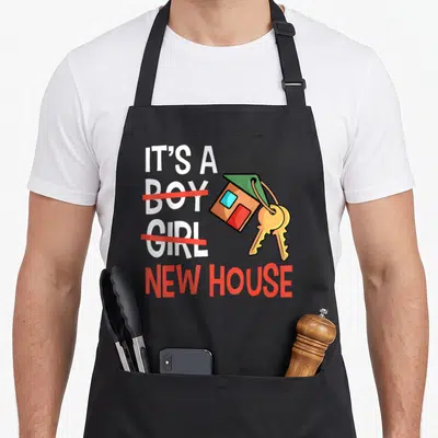 Residential housewarming apron - IT'S A BOY GIRL NEW HOUSE text with crossed out words and cartoon house keychain — shown on a black cotton canvas apron against a white background