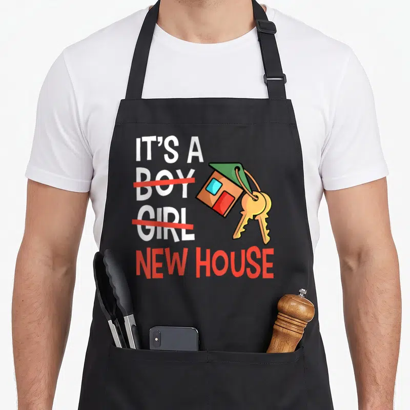 Residential housewarming apron - IT'S A BOY GIRL NEW HOUSE text with crossed out words and cartoon house keychain — shown on a black cotton canvas apron against a white background