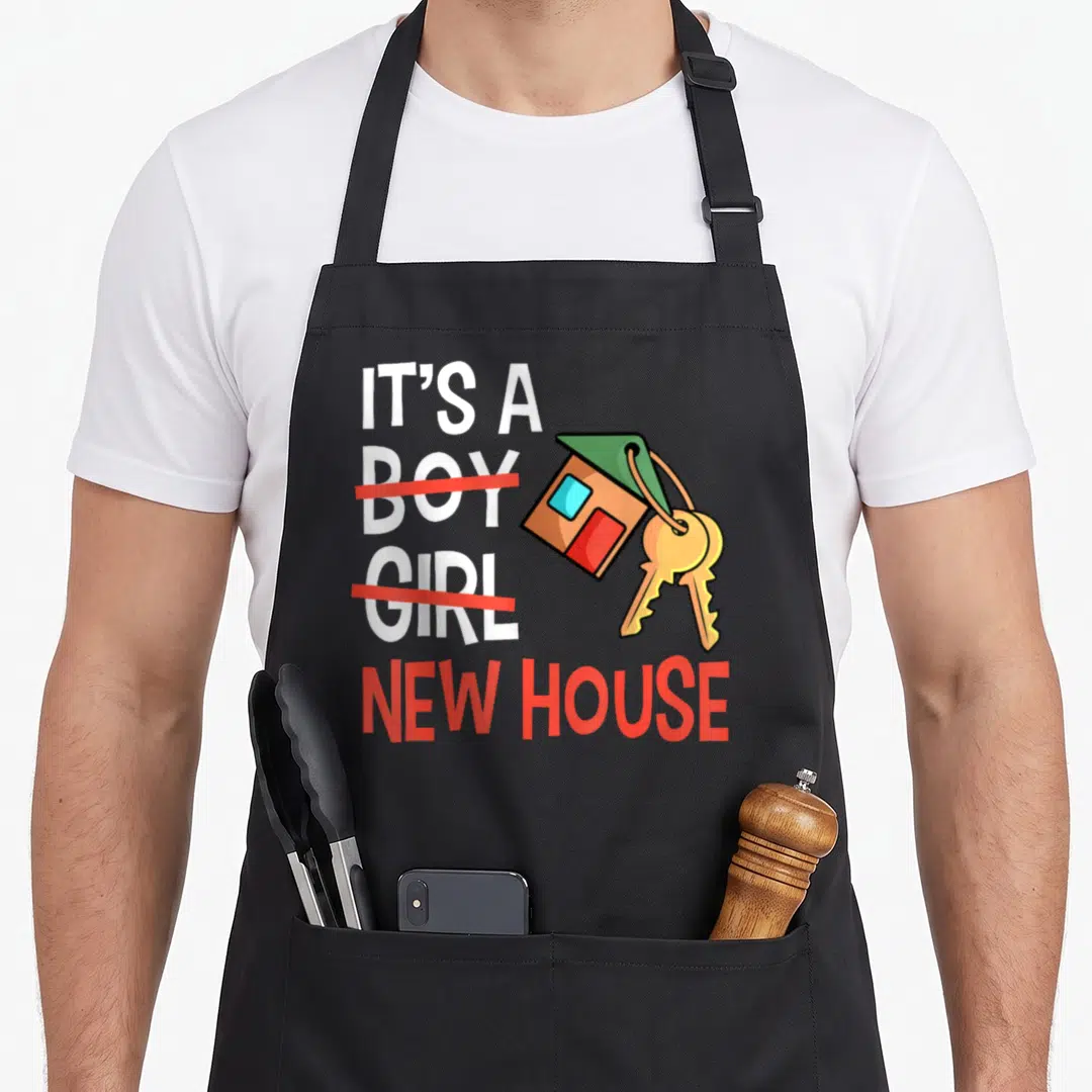 Residential housewarming apron - IT'S A BOY GIRL NEW HOUSE text with crossed out words and cartoon house keychain — shown on a black cotton canvas apron against a white background