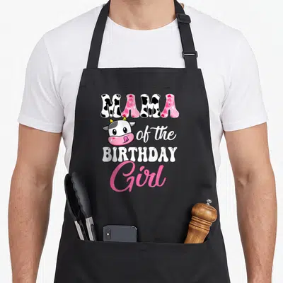 Mama farm apron - Cartoon cow head with MAMA of the BIRTHDAY Girl text in cow print and pink script — shown on a black cotton canvas apron against a white background