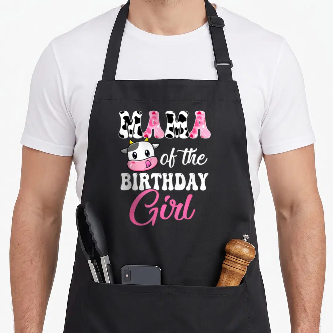 Mama farm apron - Cartoon cow head with MAMA of the BIRTHDAY Girl text in cow print and pink script — shown on a black cotton canvas apron against a white background