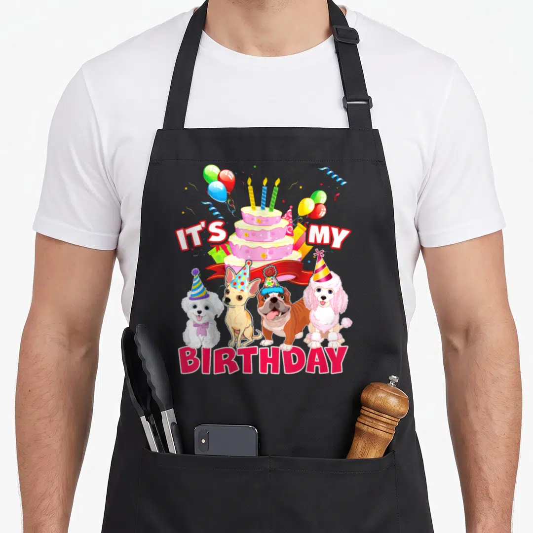 Theme dog apron - Four dogs in party hats below a tiered cake and IT'S MY BIRTHDAY text — shown on a black cotton canvas apron against a white background