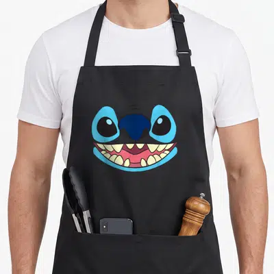 Face stitch apron - Cartoon blue face with large eyes and wide pink grinning mouth showing teeth — shown on a black cotton canvas apron against a white background