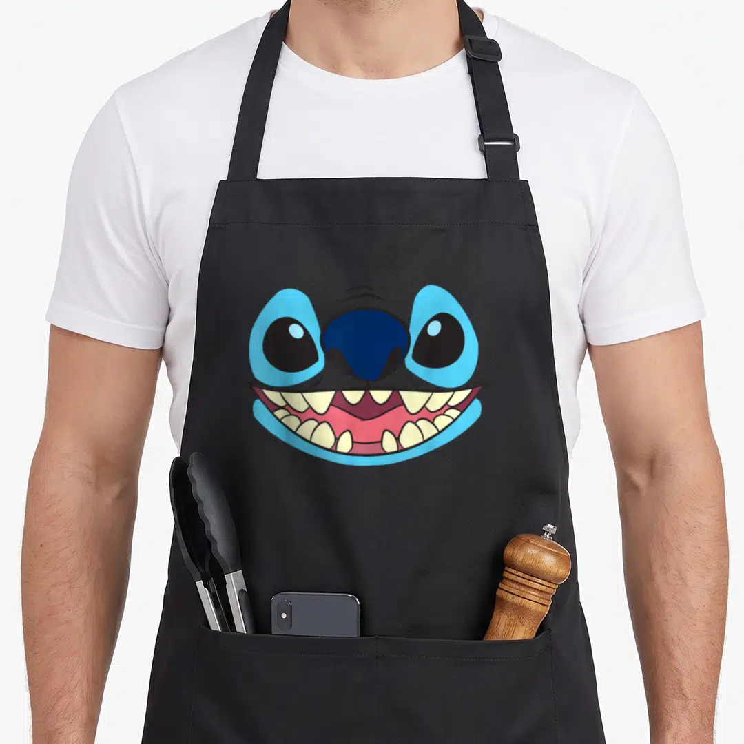 Face stitch apron - Cartoon blue face with large eyes and wide pink grinning mouth showing teeth — shown on a black cotton canvas apron against a white background