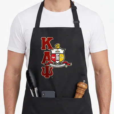 Psi alpha apron - Stacked red K A Psi letters with crest helmet shield and Greek motto ribbon — shown on a black cotton canvas apron against a white background