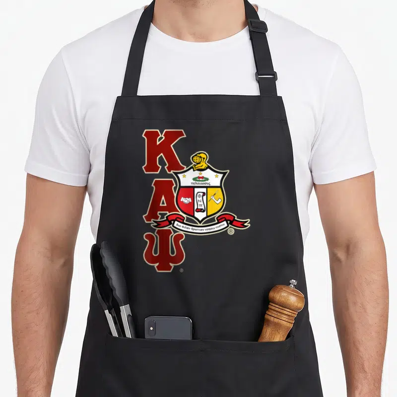 Psi alpha apron - Stacked red K A Psi letters with crest helmet shield and Greek motto ribbon — shown on a black cotton canvas apron against a white background