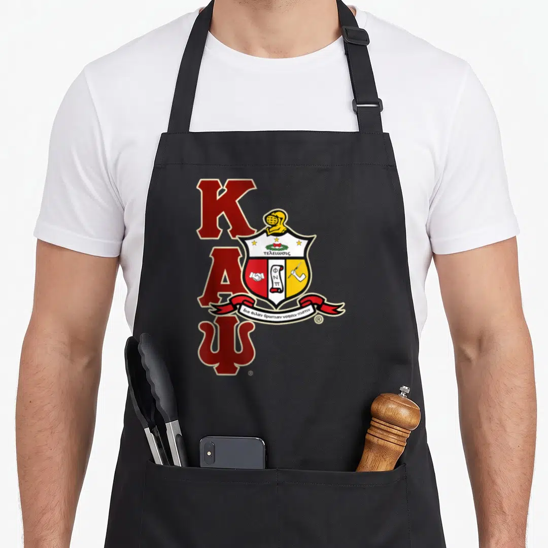 Psi alpha apron - Stacked red K A Psi letters with crest helmet shield and Greek motto ribbon — shown on a black cotton canvas apron against a white background