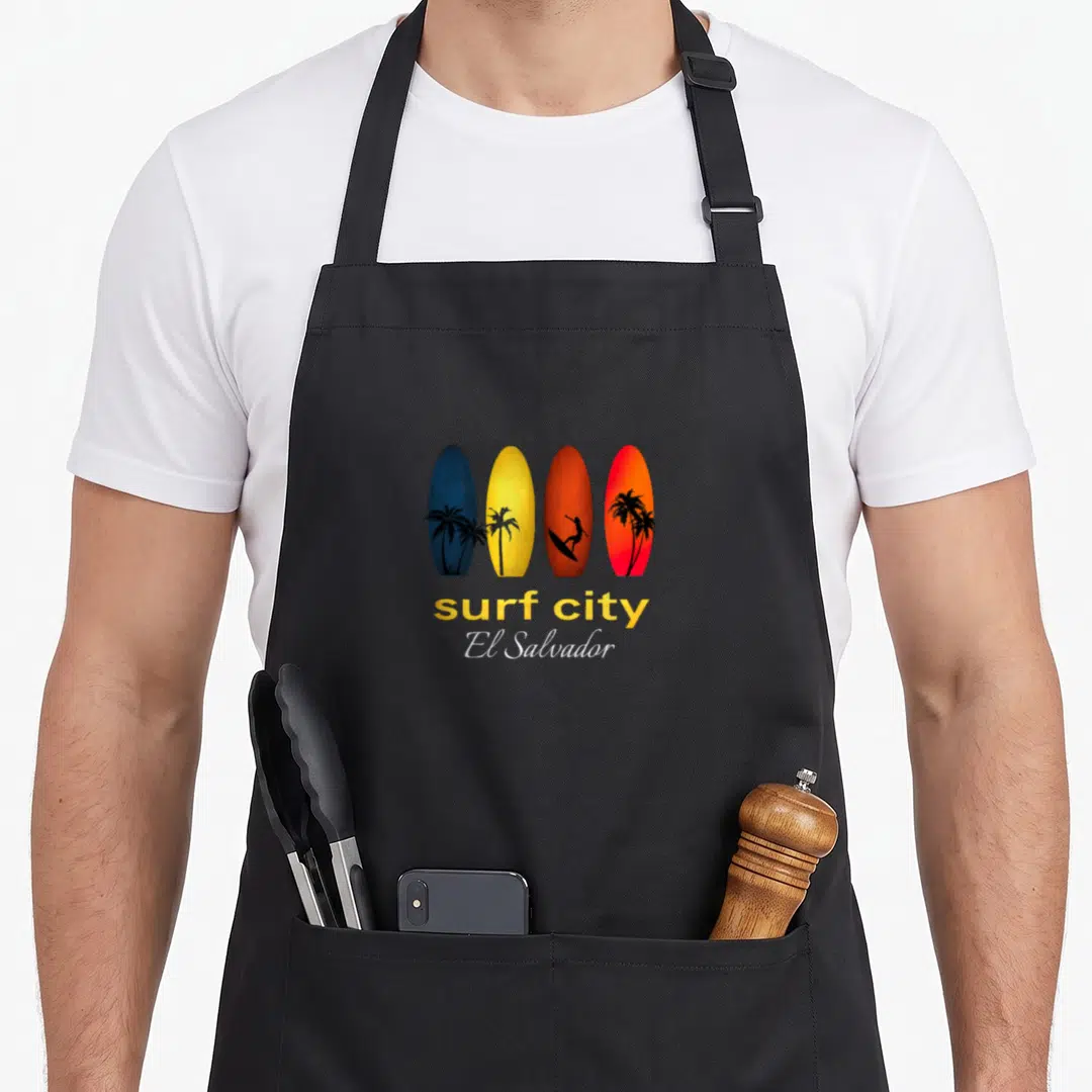 Surf el cook apron - Four colorful surfboards with palm trees and surfer silhouettes above surf city El Salvador text — shown on a black cotton canvas apron against a white background