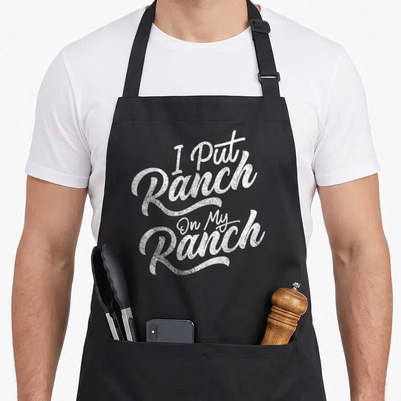 Sauce food aprons - White stacked typography reading I Put Ranch On My Ranch — shown on a black cotton canvas apron against a white background