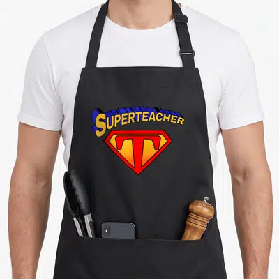 Superteacher teacher apron - Yellow and red shield with blue SUPERTEACHER text and large T inside — shown on a black cotton canvas apron against a white background