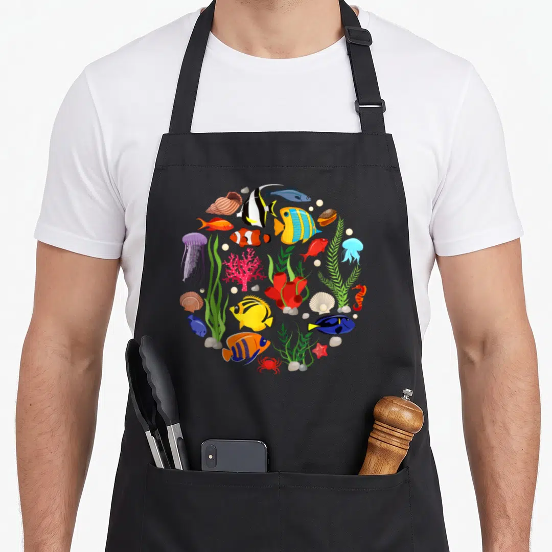 Clownfish animal apron - Cartoon circle of colorful fish, jellyfish, coral, and seashells in blue, yellow, red — shown on a black cotton canvas apron against a white background