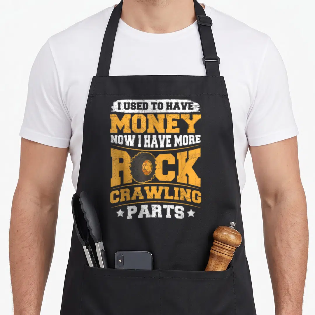 Crawling crawler girl apron - Stacked orange and white text reading I used to have money now I have more rock crawling — shown on a black cotton canvas apron against a white background