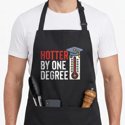 Hotter mba apron - Stacked red and white text next to a thermometer wearing a blue graduation cap — shown on a black cotton canvas apron against a white background