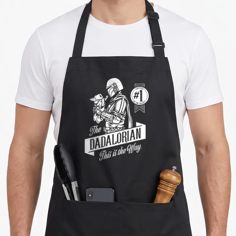 Dadalorian father day apron - White line art of Mandalorian holding Grogu with certified #1 seal and Dadalorian text — shown on a black cotton canvas apron against a white background