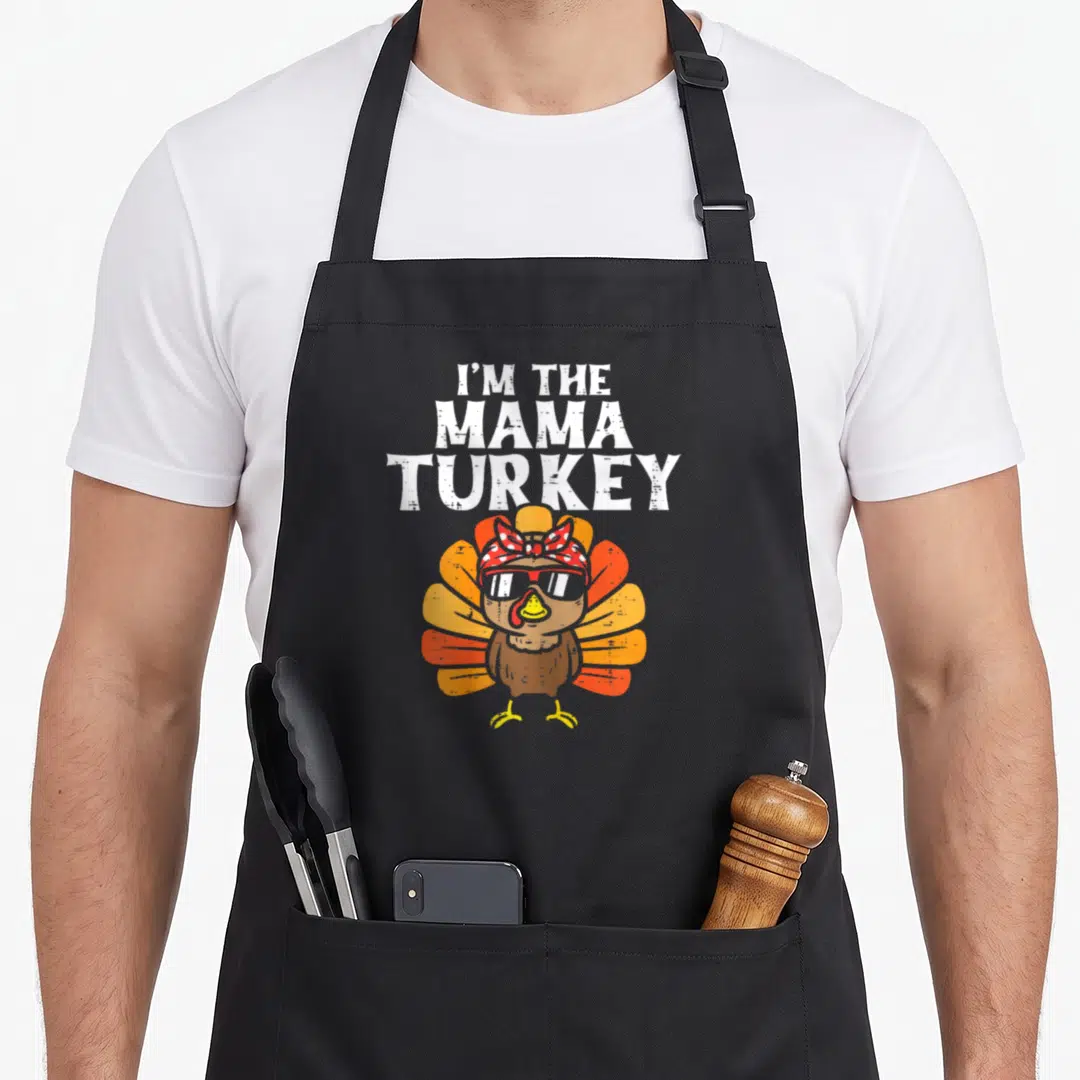 Mama thanksgiving aprons - Stacked text I'm The Mama Turkey above cartoon turkey with sunglasses and polka dot bandana — shown on a black cotton canvas apron against a white background