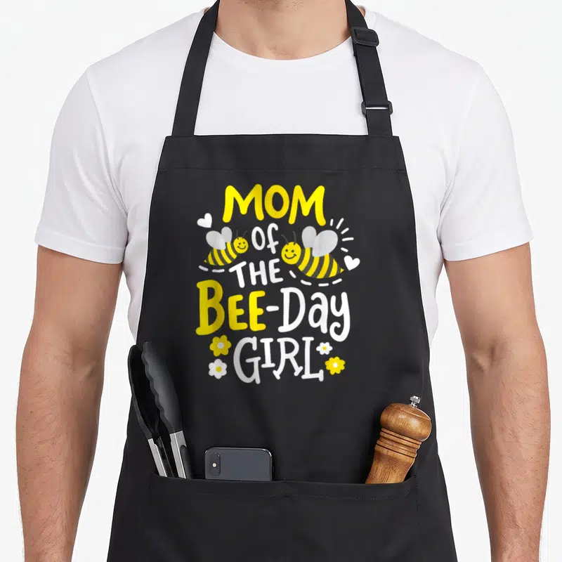 Birthday bee aprons - Stacked MOM Of THE BEE-Day GIRL text with two flying bees and small hearts flowers in yellow — shown on a black cotton canvas apron against a white background