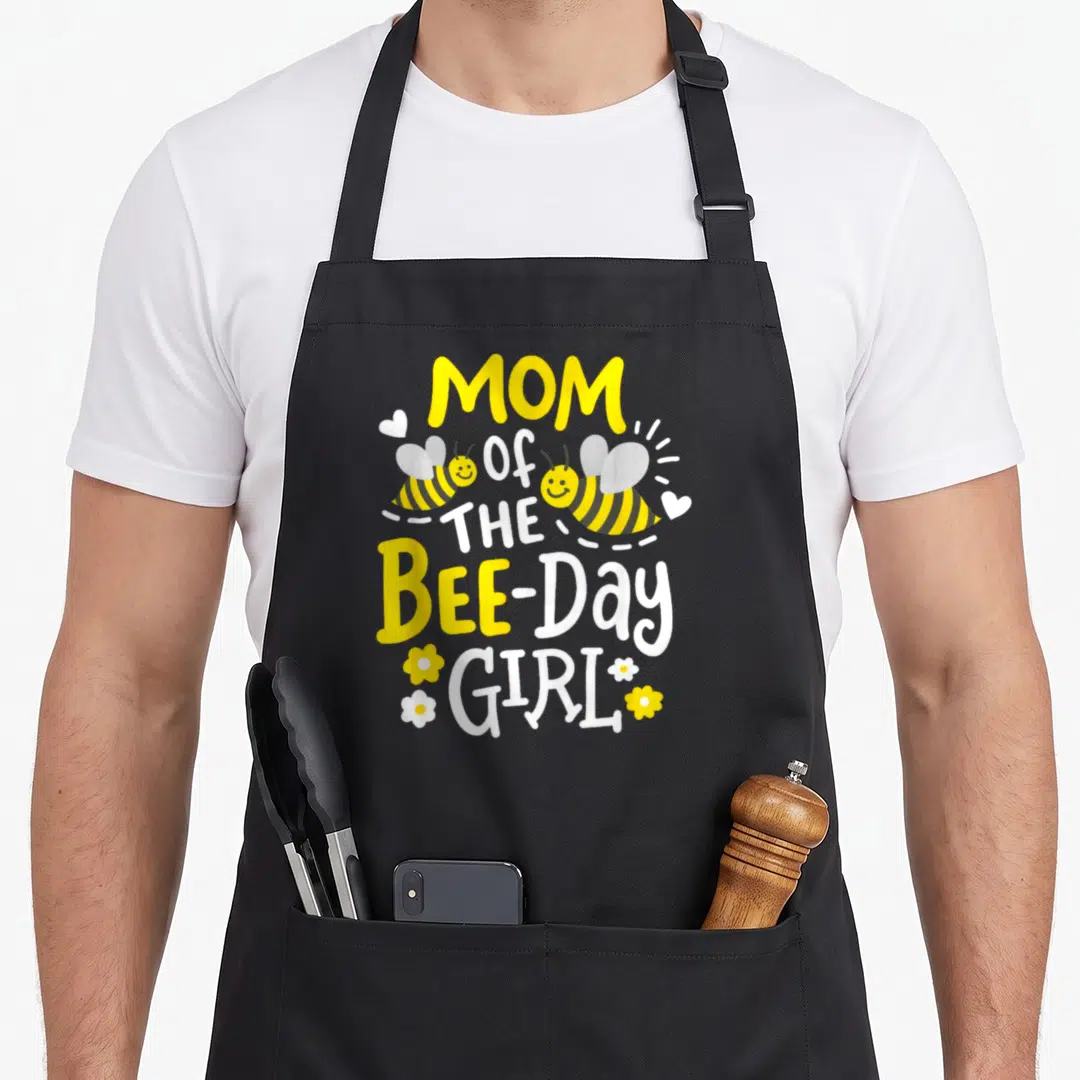 Birthday bee aprons - Stacked MOM Of THE BEE-Day GIRL text with two flying bees and small hearts flowers in yellow — shown on a black cotton canvas apron against a white background
