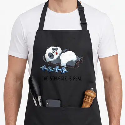 Panda bodybuilding apron - Cartoon panda lying next to blue barbell with text THE STRUGGLE IS REAL — shown on a black cotton canvas apron against a white background