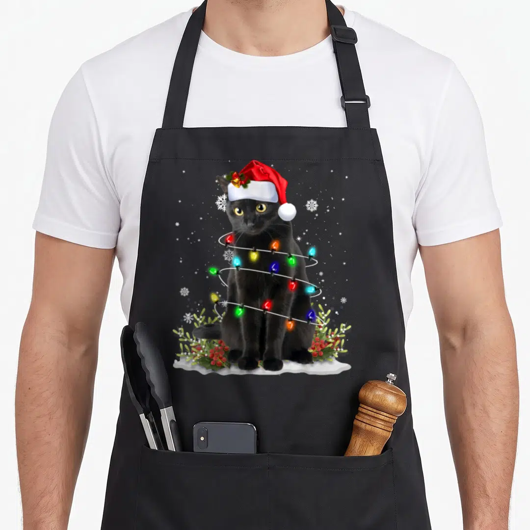 Cat santa aprons - Black cat in red Santa hat wrapped in colorful lights with holly and snowflakes — shown on a black cotton canvas apron against a white background