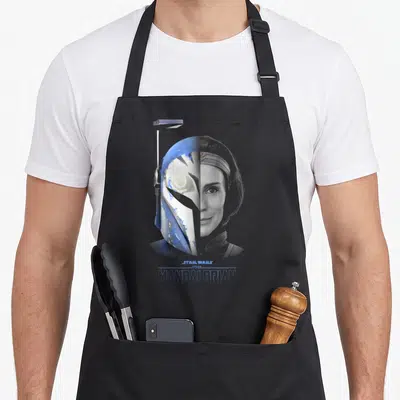 Katan split apron - Split face design with blue helmet on left and grey female face on right, centered text below — shown on a black cotton canvas apron against a white background