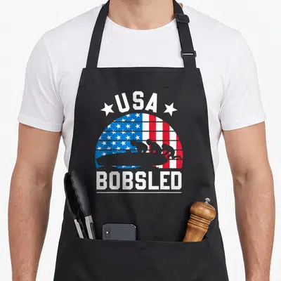 Bobsled bobsledding american flag apron - USA BOBSLED text with grey bobsled team silhouette over red white blue flag — shown on a black cotton canvas apron against a white background