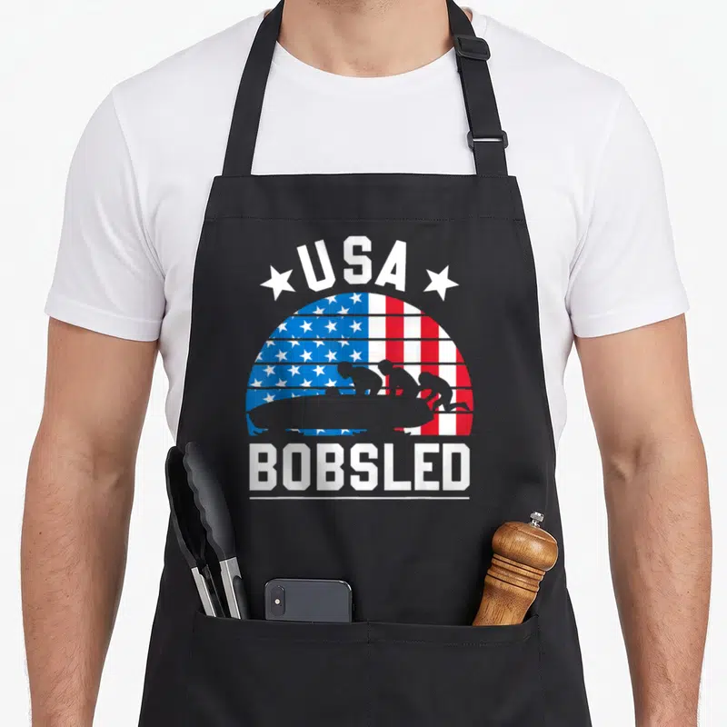 Bobsled bobsledding american flag apron - USA BOBSLED text with grey bobsled team silhouette over red white blue flag — shown on a black cotton canvas apron against a white background