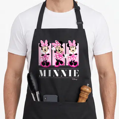 Grid friends apron - Three Minnie Mouse figures in pink dresses above white MINNIE text between horizontal lines — shown on a black cotton canvas apron against a white background