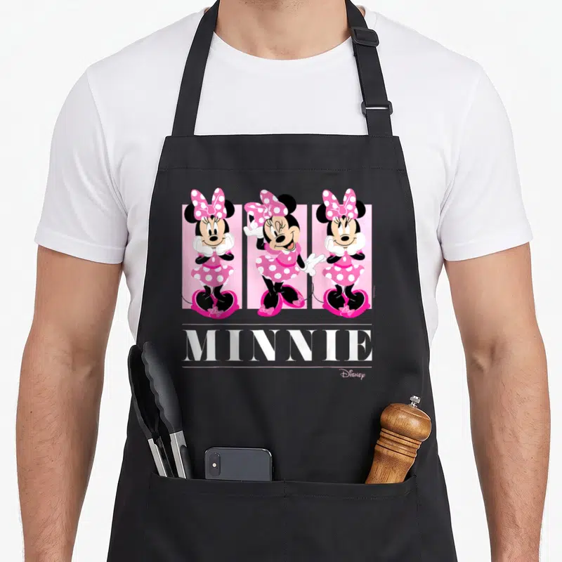 Grid friends apron - Three Minnie Mouse figures in pink dresses above white MINNIE text between horizontal lines — shown on a black cotton canvas apron against a white background