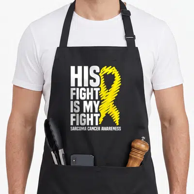 His sarcoma awareness apron - White stacked text His Fight Is My Fight next to yellow scribbled ribbon on black apron — shown on a black cotton canvas apron against a white background