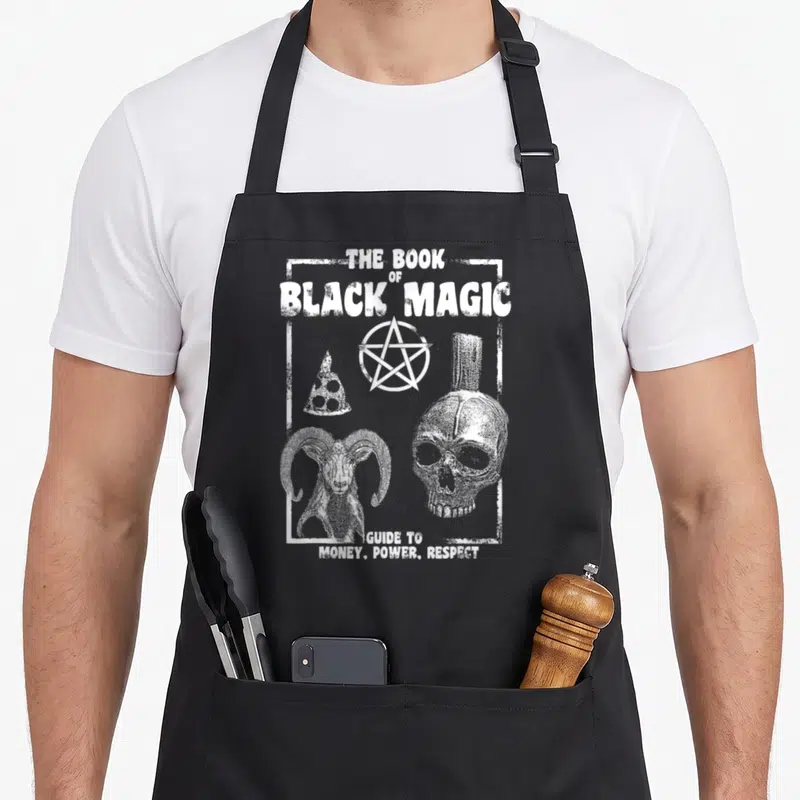 Alt gothic apron - White distressed text The Book of Black Magic Guide to Money Power Respect with skull pentagram — shown on a black cotton canvas apron against a white background