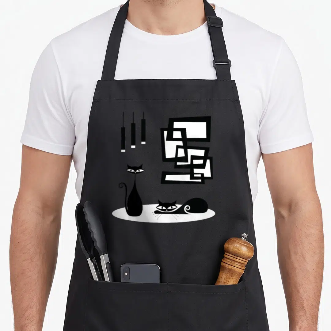 Machine modern apron - Two black cats on white rug with hanging lamps and geometric frames — shown on a black cotton canvas apron against a white background