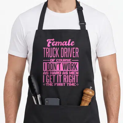 Right truck apron - Pink stacked typography reading Female TRUCK DRIVER OF COURSE I DON'T WORK AS HARD AS MEN I GET IT — shown on a black cotton canvas apron against a white background