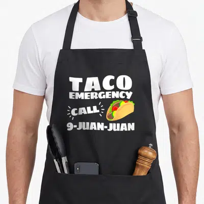 Cinco taco apron - TACO EMERGENCY CALL 9-JUAN-JUAN text next to cartoon taco with lettuce and tomato — shown on a black cotton canvas apron against a white background